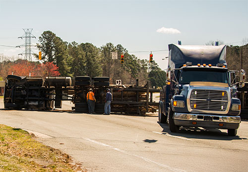 An overturned semi-truck is lying on the side of a road while another one approaches.