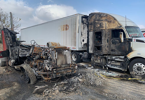 The charred remains of a semi-truck are on the side of a road after a collision.